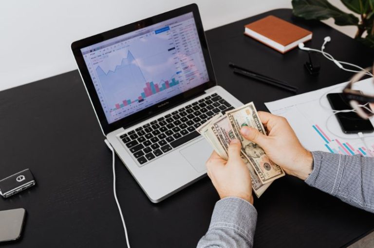Pessoa realizando conciliação bancária com análise de dados financeiros no notebook e conferência de valores em dinheiro.